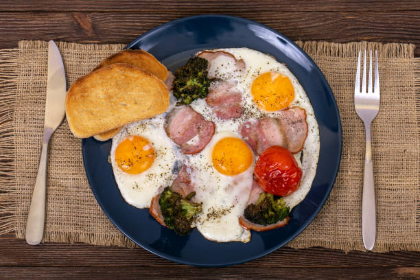 istock Delicious breakfast with fried eggs, bacon, tomato, broccoli and toasts in the plate that is on burlap, on wooden background 1264649027