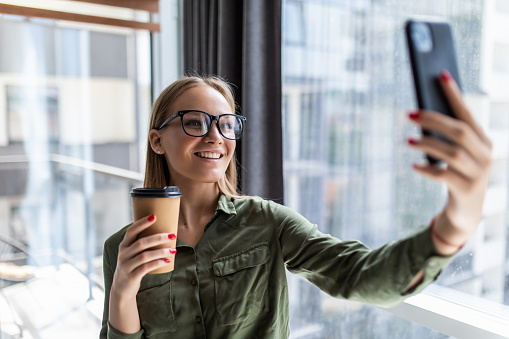 Photo of woman in formal wear standing holding takeaway coffee in hand and taking selfie on mobile phone in office