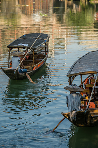 Feng Huang, China -  August 2019 : Chinese men wearing traditional conical wicker hats sitting in a long narrow empty wooden tourist boats  on the Tuo river, flowing through the centre of Fenghuang Old Town