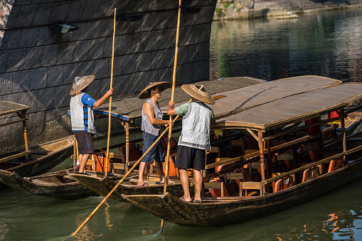 Feng Huang, China -  August 2019 : Three Chinese men wearing traditional conical wicker hats sitting in a long narrow empty wooden tourist boats  on the Tuo river, flowing through the centre of Fenghuang Old Town