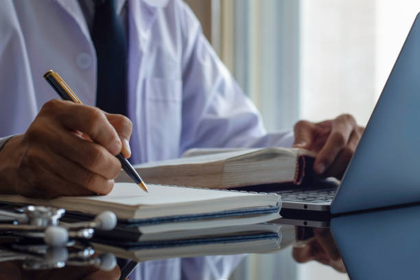 Doctor Closeup male doctor or physician reading text book and writing information,work on laptop computer with medical stethoscope on the desk. Online working, telehealth or meditech concept. medical journal stock pictures, royalty-free photos & images