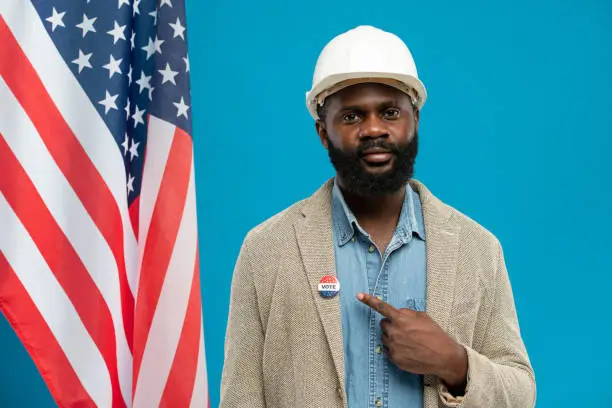 Bearded engineer of African ethnicity standing against stars-and-stripes flag and pointing at vote insignia on collar of his jacket
