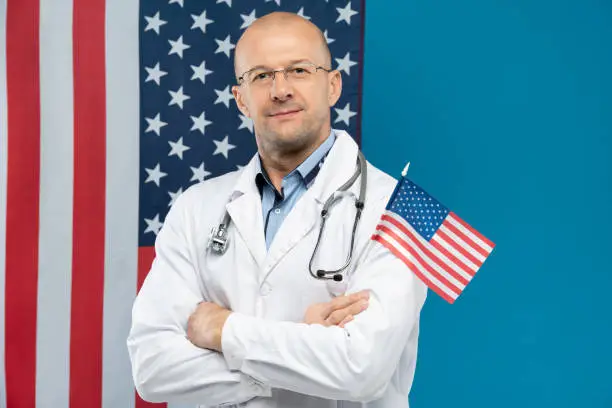 Bald doctor in whitecoat and eyeglasses holding small stars-and-stripes while standing in front of camera against wall with American flag