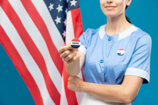 Young room-maid in blue uniform showing you vote insignia while standing against stars-and-stripes flag background