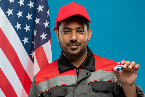 Young serious repairman of Hispanic ethnicity holding vote insignia against stars-and-stripes flag while standing in front of camera