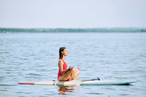 giovane sportiva in costume da bagno rosso che pratica yoga mentre è seduta sulla tavola da surf - paddleboard oar women lake foto e immagini stock