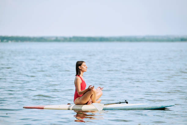 young sportswoman in red swimsuit practicing yoga while sitting on surfboard - women paddleboard bikini surfing imagens e fotografias de stock