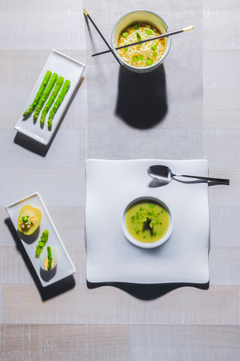Top view of a vegetable soup dish surrounded by a bowl and chopsticks, a plate with asparagus and another with potato, egg and chopped vegetables. Healthy food concept.