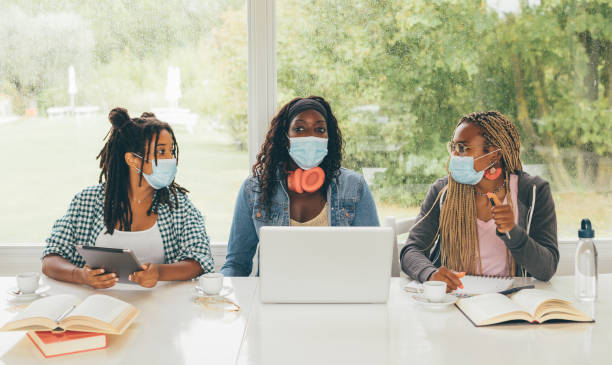 Back to school concept. Three African American girls students sitting at the table preparing for exam or making homework together, they are using lap top and digital tablet wearing surgical masks. Back to school concept. college students studying together stock pictures, royalty-free photos & images