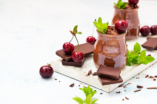 Chocolate mousse with mint and sweet cherry in portion glasses on wooden cutting board. Light blue concrete background.