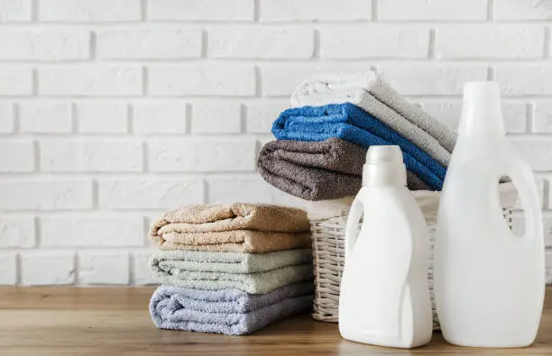 Photo of Laundry concept. Bottles with detergent gel and lush towels in in basket on wooden table with white brick wall on background.