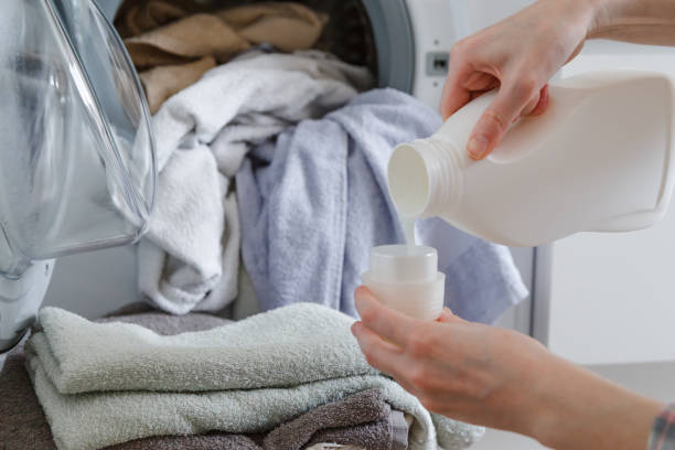 Close up of female hands pouring liquid laundry detergent into cap. Washer machine and clothes with wicker basket in background stock photo