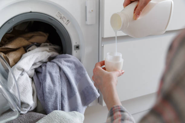 istock Close up of female hands pouring liquid laundry detergent into cap on white rustic table with towels on background in bathroom. 1264705924