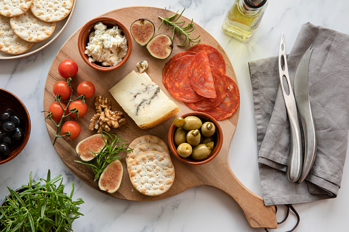Truffle Pecorino cheese, Gorgonzola, cherry tomatoes, green olives with jalapeños, figs, savoury biscuits, crackers, olive oil, rosemary pot, cheese knives, black olives, walnuts, grey napkin, on a round timber wooden board, platter.