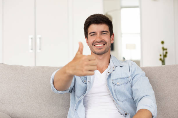 istock Happy guy smiling at camera and shows thumb up, sitting on sofa at home. 1264718587