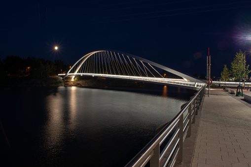 An illuminated walking bridge casting a reflection on the water during night time. The bridge connects new residential area of Kalasatama and Helsinki Zoo Korkeasaari.