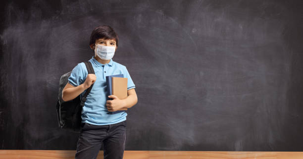 niño en la escuela con una mochila y libros de pie frente a una pizarra de la escuela y usando una máscara facial - little boys measuring expressing positivity intelligence fotografías e imágenes de stock