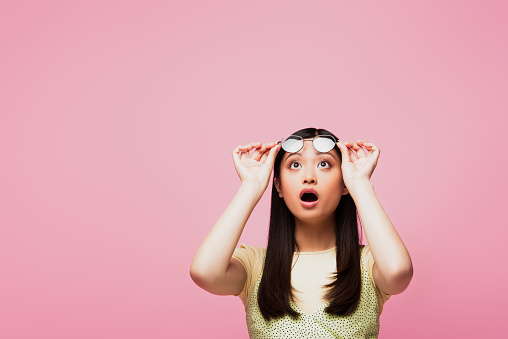 shocked asian girl looking up and touching glasses isolated on pink
