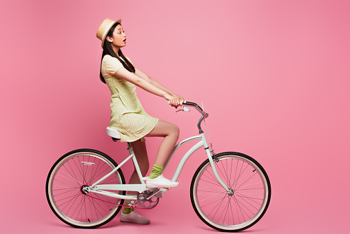 side view of shocked asian young woman in straw hat riding bicycle on pink