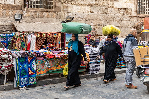 Egyptian women in traditional clothing, carrying shopping bags on top of their heads, while walking through the rug market in the historic Khan el-Khalili souk, Cairo.