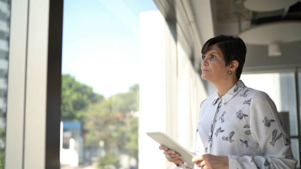 istock Businesswoman looking through window contemplating and using digital tablet on the office 1264739512