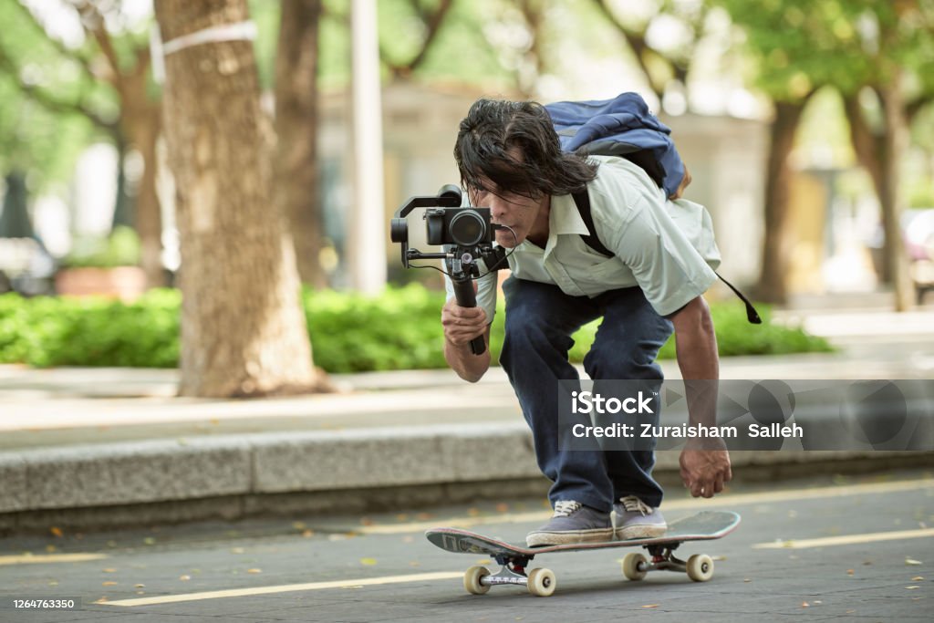 Asian skateboarder filming using video recorder with gimbal Asian young man with skateboard shooting video with gimbal Camera Operator Stock Photo