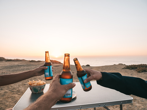 Three friends holding up beers cheering at sunset while sitting on camping chairs