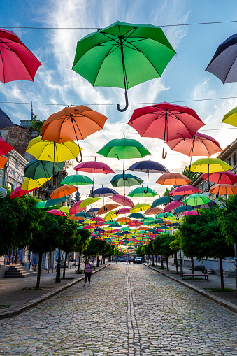 Braila, Romania - 25 July, 2020: wide angle color image depicting a street scene on the elegant cobblestone streets of old town Braila, Romania. People are walking past, going about their daily business, while overhead rows of multi colored umbrellas recede into the distance.