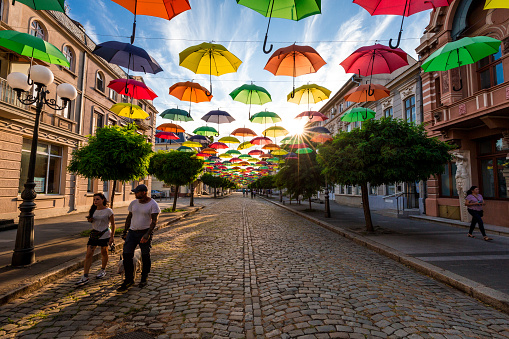 Braila, Romania - 25 July, 2020: wide angle color image depicting a street scene on the elegant cobblestone streets of old town Braila, Romania. People are walking past, going about their daily business, while overhead rows of multi colored umbrellas recede into the distance.