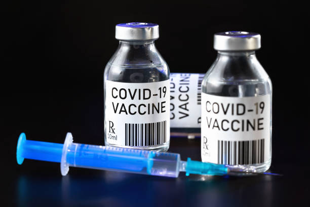 istock Coronavirus Covid-19 vaccine concept -  three glass vials on black table, hypodermic syringe needle near, closeup detail (label own design - dummy data and barcode, not real product) 1264808013