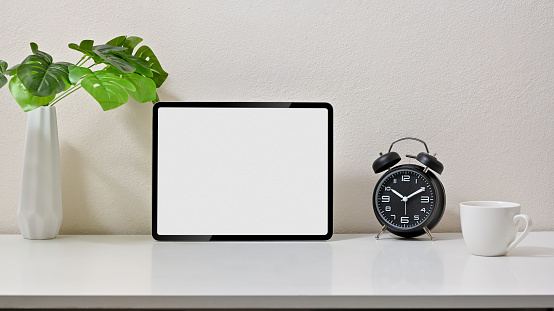 Mock up stylish of workspace with blank screen tablet, clock, coffee cup and vase on white table and white background. Front view.