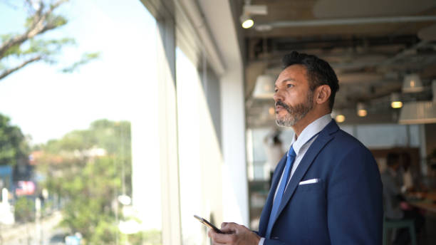 Businessman looking through window contemplating and using mobile phone on the office