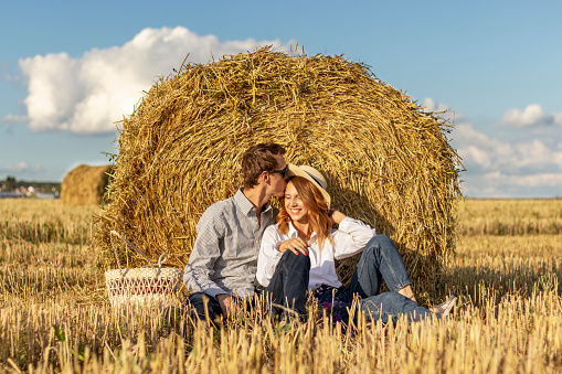 Beautiful young couple in love in casual clothes sitting in field near hay bale. Ecotourism in countryside concept.