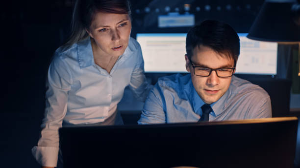 istock Portrait of Businessman Uses Desktop Computer, His Female Project Manager Explains Specific Tasks, Account Handling and Strategic Moves. Professional People Late at Night in Big Corporate Office 1264819503