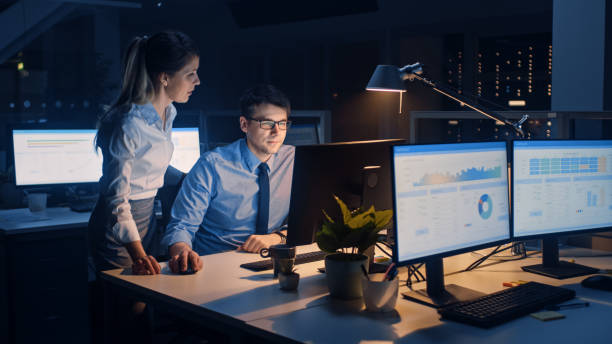 istock Businessman Uses Desktop Computer, His Female Project Manager Explains Specific Tasks, Account Handling and Strategic Moves. Professional People Late at Night in Big Corporate Office 1264819522