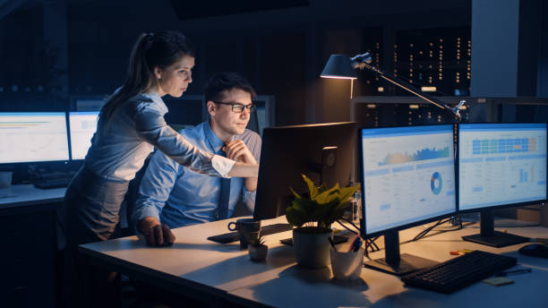 istock Businessman Uses Desktop Computer, His Female Project Manager Explains Specific Tasks, Account Handling and Strategic Moves. Professional People Late at Night in Big Corporate Office 1264819523