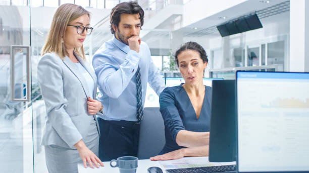 istock In Bright Modern Office: Office Worker Sitting and Working at Her Desktop Computer, Discusses Problem Solution with Her Project Manager and Team Leader. Young Businessmen Planning and Problem Solving 1264819595