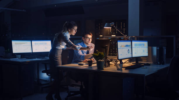 istock Businessman Uses Desktop Computer, His Female Project Manager Explains Specific Tasks, Account Handling and Strategic Moves. Professional People Late at Night in Big Corporate Office 1264819602