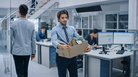 Sad Fired / Let Go Office Worker Packs His Belongings into Cardboard Box and Leaves Office. Workforce Reduction, Downsizing, Reorganization, Restructuring, Outsourcing. Shot with Dark Ambient