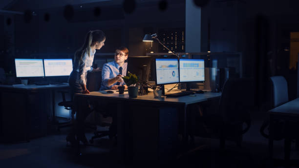 istock Ambitious and Confident Businessman Uses Desktop Computer, His Female Project Manager Explains Specific Tasks, Account Handling and Strategic Moves. Professional People Late at Night in Big Office 1264819645