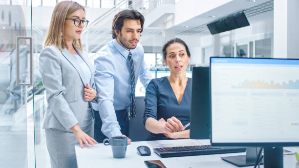istock In Bright Modern Office: Office Worker Sitting and Working at Her Desktop Computer, Discusses Problem Solution with Her Project Manager and Team Leader. Young Businessmen Planning and Problem Solving 1264819673