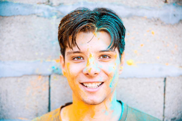 Young man face covered with Holi colors for Indian traditional festival on the street. Colorful handsome Man happy face looking at camera. Guy with holi color powder on head outdoors