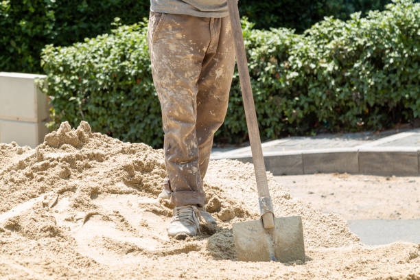 Construction worker in dirty uniform holding in his hand a shovel standing on a pile of sand and is about to shovel sand, Digging a pit by shovel. Construction outside in the street, roadworks