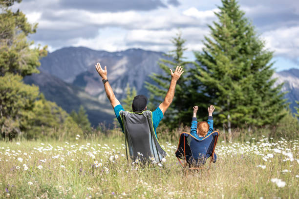 istock Father and Son sitting on camping chairs with arm raised and looking at mountains 1264836366
