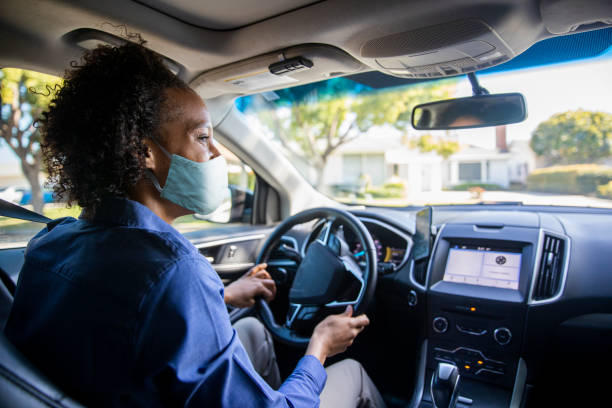 Young Black Woman Driving Car for Rideshare Wearing a Mask A young black woman drives a passenger in her car as a professional driver while wearing a mask. car pooling photos stock pictures, royalty-free photos & images