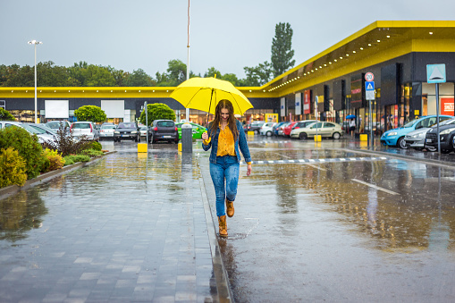 Young woman with yellow umbrella jumping in the water puddles on a rainy day.