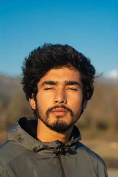 headshot of a handsome 18-20 years old man with his eyes closed in a winter evening/morning. background contains trees, mountains and clear blue sky. background is blurred. his bearded face is sunlit.