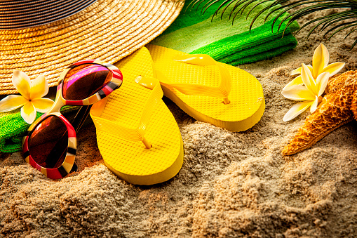 This is a photograph of sandals, sunglasses, a hat and a palm branch on a sandy beach