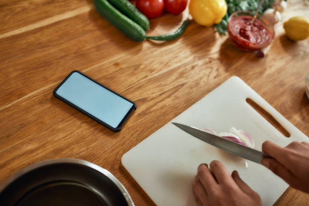 istock Close up of phone with blank screen on the table. Man cutting onion on chopping board, using smartphone app while cooking dinner. Healthy nutrition, technology concept 1264886536