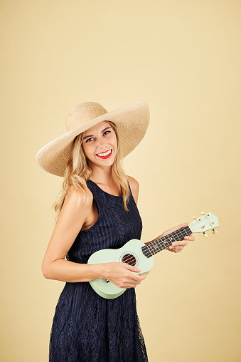 Portrait of smiling pretty blond young woman in straw hat playing ukulele, isolated on yellow